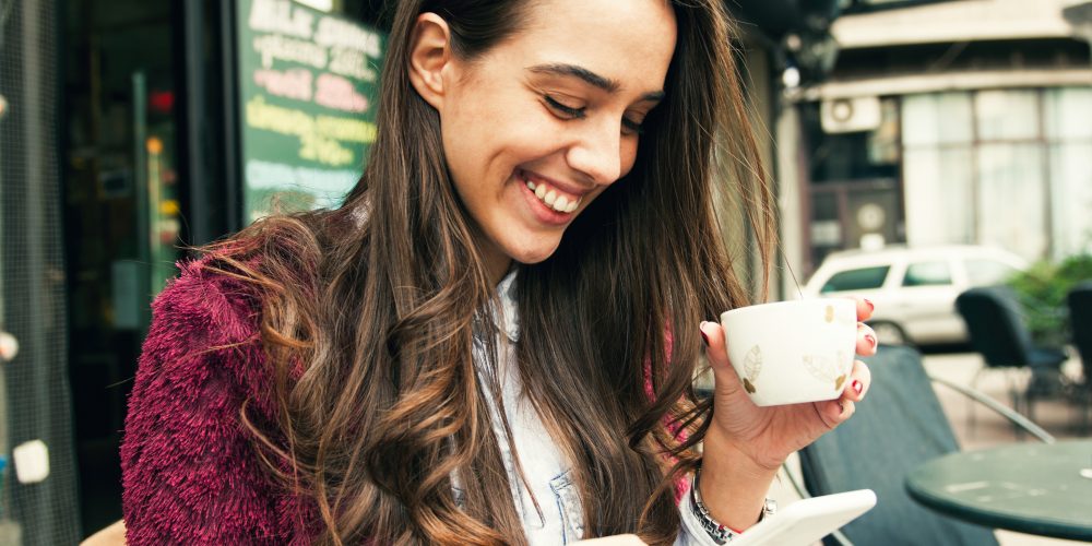 Young smiling woman using smart phone while drinking coffee