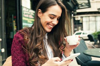 Young smiling woman using smart phone while drinking coffee