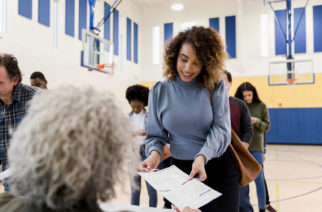 The unrecognizable mature adult female polling place volunteer assists the mid adult woman with the document she needs to be aloe to vote.  Other voters use smart phones while waiting in line.
